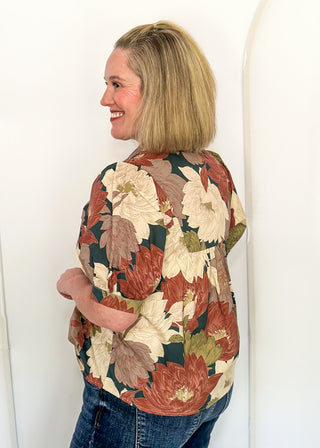 A woman with blonde hair is shown from the back wearing a short-sleeve button-up shirt with a large floral pattern in rust red, cream, and olive green, paired with blue jeans, standing against a white arched wall.