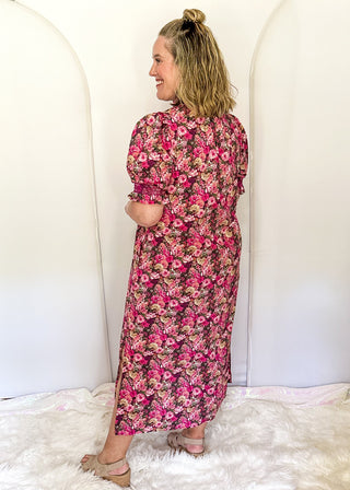 A woman stands with her back to the camera wearing a pink and red floral midi dress with puff sleeves, posing on a white faux fur rug in front of white arch backdrops.