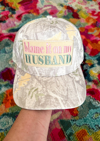 A camouflage baseball cap held by a hand, featuring a front patch that reads 'blame it on my HUSBAND' in pink and teal lettering, set against a colorful patterned rug.