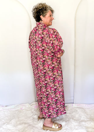 An older woman stands in profile wearing a long-sleeve, knee-length floral dress with a pink, red, and beige pattern on a dark background. She is standing on a white faux fur rug in front of a white arched backdrop.
