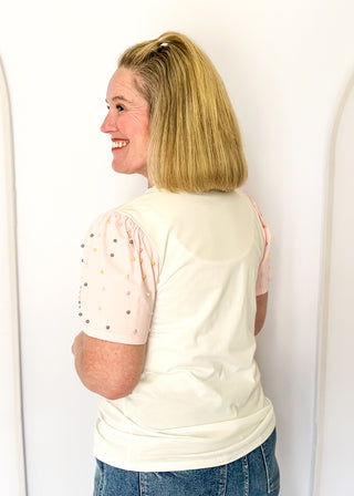 Off white short sleeve shirt with colorful embellished text and pink statement sleeves with decorative pearl studs.