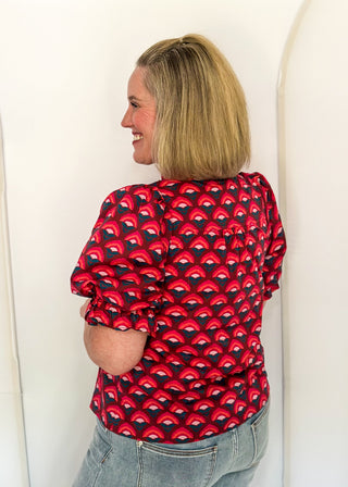 A woman with blonde hair is shown from the back wearing a red short-sleeve top with a black and red repeating scallop pattern, paired with light blue jeans, standing against a white arched wall.