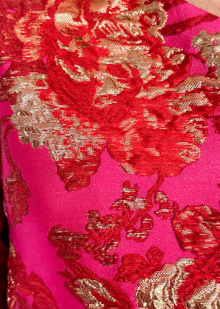A close-up of a vibrant magenta pink fabric with an intricate red and metallic gold floral brocade pattern.