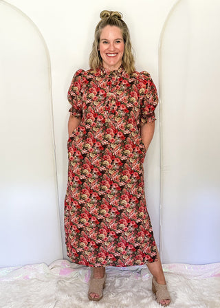 A woman stands smiling in a floral midi dress with puff sleeves, standing on a white faux fur rug in front of a white arched backdrop.