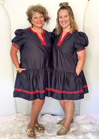 Two women wearing matching navy dresses with red trim standing against a white background.