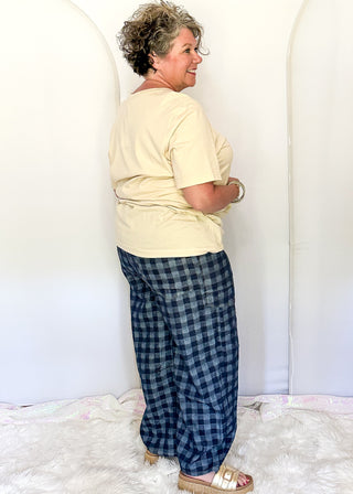 A woman wearing a cream short-sleeve t-shirt and blue-and-black plaid pants, standing in profile against a white studio backdrop with arched panels and a white faux-fur rug.