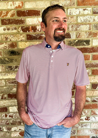 A man wearing a pink and white striped polo shirt with blue jeans, standing in front of a rustic brick wall.