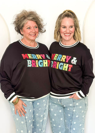 Two women standing side-by-side wearing matching black sweatshirts with 'MERRY & MERRY BRIGHT' printed in colorful letters across the chest, paired with light blue polka dot jeans.