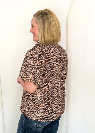 A woman with blonde hair is shown from the back wearing a leopard print short-sleeve button-up shirt and blue jeans, standing against a white studio backdrop with arched panels.