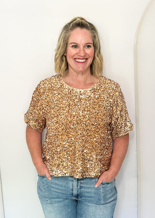 A smiling blonde woman wearing a short-sleeve gold sequined top and blue jeans, standing against a white studio backdrop with curved white panels.
