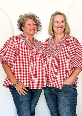 Two women standing side by side in matching red and white gingham short-sleeve button-up shirts and blue jeans, posing in front of a white photo backdrop with a ceiling fan visible above.