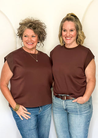Two women standing side-by-side wearing matching brown short-sleeve crewneck tops and blue jeans, posing in front of a white draped studio backdrop.
