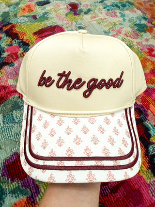 A cream-colored baseball cap with 'be the good' embroidered in dark red script on the front. The brim features a white background with a repeating red damask pattern and dark red piping.
