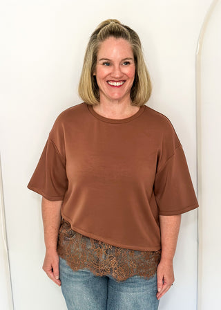 A smiling woman with blonde hair stands against a white arched wall, wearing a brown short-sleeve crewneck top, a brown lace hem detail, and blue jeans.