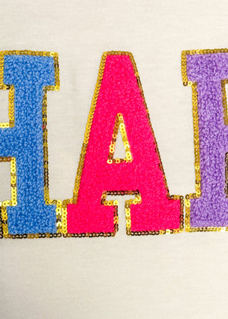 Close up of lightweight cream long-sleeve top with colorful chenille patch letters spelling “HAPPY”