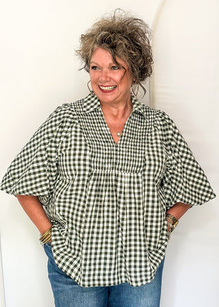 A smiling woman with curly gray hair stands against a white arched backdrop, wearing a black and white gingham short-sleeve blouse with blue jeans.