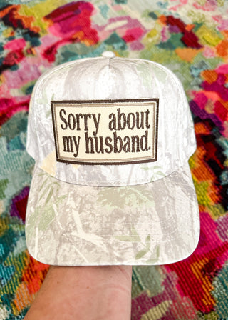 A white camouflage baseball cap held by a hand, featuring a rectangular embroidered patch on the front that reads 'Sorry about my husband.' in brown text on a beige background.