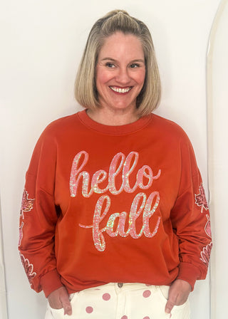 woman wearing an orange long sleeve top with sequin leaves on the sleeves and "hello fall" across the front in sequins.
