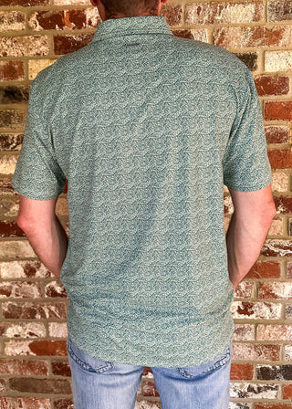 A man viewed from behind wearing a short-sleeve collared button-up shirt with a green and white abstract pattern, paired with light blue jeans, standing against a brick wall.