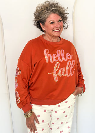 woman wearing an orange long sleeve top with sequin leaves on the sleeves and "hello fall" across the front in sequins.