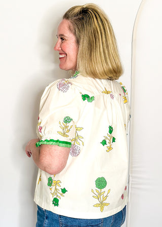 off white floral short sleeve button front blouse with pink and green flowers, featuring puff sleeves, ruffled neckline, and green trim.