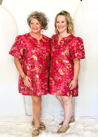 Two women standing side by side wearing matching short puff-sleeve dresses in a vibrant pink and gold floral jacquard fabric, posed against a white circular backdrop.