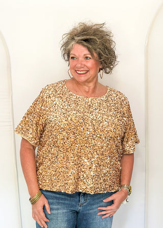 A woman with short, curly gray hair wearing a gold sequined short-sleeve top and blue jeans, standing against a white arched backdrop.
