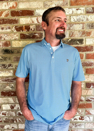 A man wearing a light blue polo shirt and jeans stands against a rustic brick wall, smiling and looking to the side.