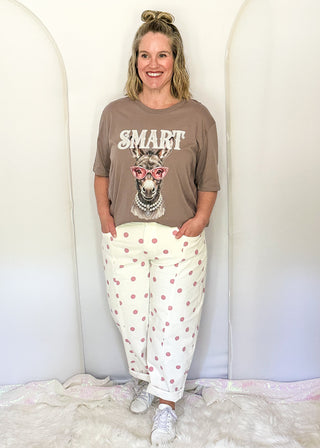 A woman stands indoors wearing a brown graphic T-shirt with a llama illustration and the word 'SMART' across the front, paired with cream pants covered in pink polka dots, white sneakers, and a white faux-fur rug against a white arched backdrop.