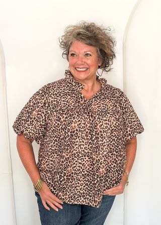A smiling woman with curly gray hair wearing a brown and beige leopard-print short-sleeve blouse and blue jeans, standing against a white arched wall.