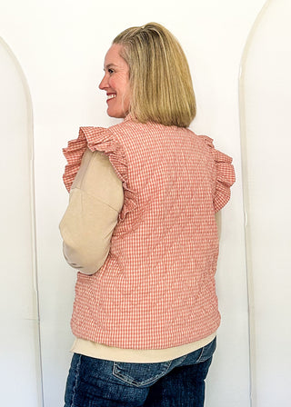 A woman with blonde hair is shown from the back, wearing a pink and white gingham ruffled sleeveless vest over a beige long-sleeve shirt and dark blue jeans, standing in front of a white arched backdrop.