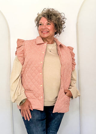 A smiling woman with curly gray hair stands in front of white arched panels, wearing a pink and white gingham ruffled sleeveless vest over a beige long-sleeve top and blue jeans.