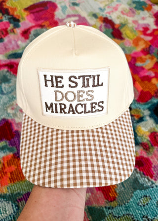 A cream-colored baseball cap with a brown and white gingham patterned brim. The front has a white patch with the embroidered text 'HE STIL DOES MIRACLES' in brown.