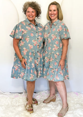 Two women standing on a white carpet wearing button front denim dresses with pink floral design.  