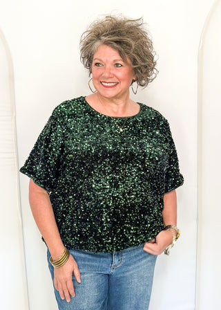 A smiling woman with short curly gray hair models a dark green sequined short-sleeve top and blue jeans against a white arched backdrop.