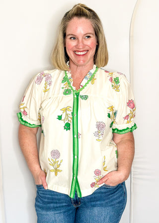 off white floral short sleeve button front blouse with pink and green flowers, featuring puff sleeves, ruffled neckline, and green trim.