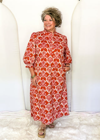 A woman modeling a red and white patterned midi dress with puff sleeves, standing on a white faux fur rug in front of white arched backdrop panels.
