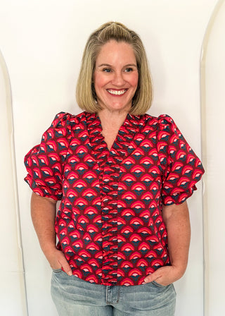 A smiling woman with blonde hair stands against a white arched wall, wearing a red and black patterned short-sleeve blouse with ruffles and light-wash blue jeans.
