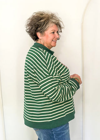 An older woman with short gray hair is shown from the side, wearing a green and white striped long-sleeve knit top and blue jeans, standing against a white studio backdrop with arched panels.
