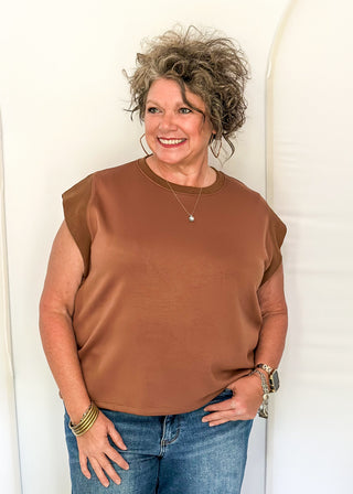 A smiling woman with curly gray hair wearing a brown short-sleeve top and blue jeans, standing against a white arched backdrop.