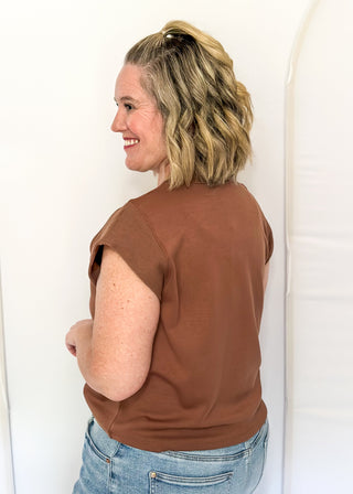 A woman with blonde hair tied back is shown from the back wearing a brown short-sleeve top and light blue jeans, standing against a white studio backdrop.