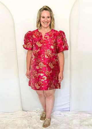 A woman stands in a studio wearing a short puff-sleeve dress with a bright pink and gold floral pattern, paired with gold flat shoes, against a white arched backdrop.