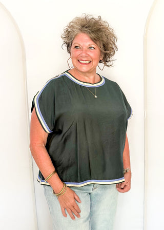 A smiling woman with curly gray hair wearing a dark green short-sleeve top with white and blue striped trim, paired with light blue jeans, standing against a white arched backdrop.