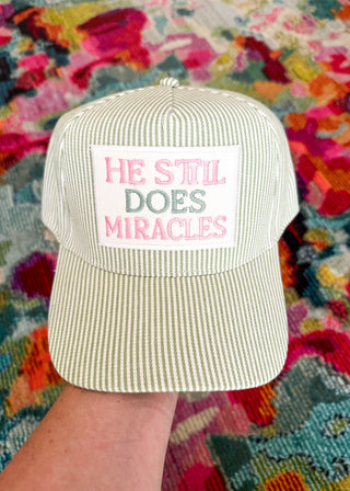 A light beige and white vertically striped baseball cap held by a hand, featuring a white front patch with the embroidered text 'HE STIL DOES MIRACLES' in pink and green. The background is a highly colorful, textured patterned rug.