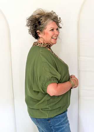 A woman with short curly gray hair models an olive green short-sleeve top with a leopard-print collar, paired with blue jeans, standing against a white arched backdrop.