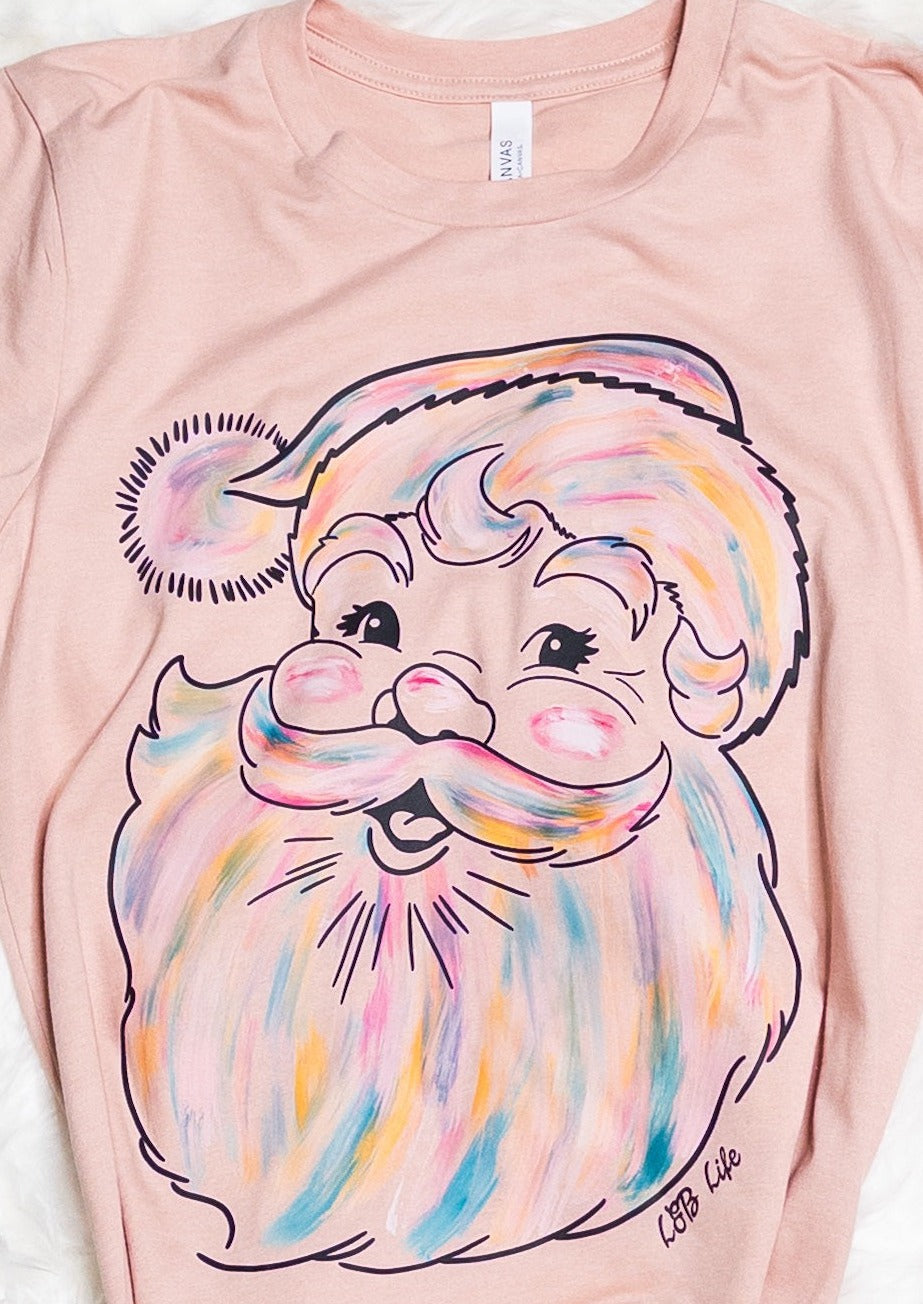 Peach colored tee shirt with an image of Santa's face. It is mostly the color of the tee shirt but has a brushstroke look of many colors, including pink, blue, white, yellow, and purple.