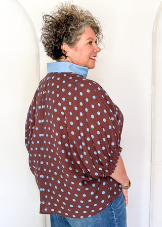 A woman wearing a brown long-sleeve blouse with light blue polka dots and a light blue collar, paired with blue jeans, standing against a white arched wall.