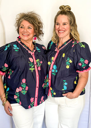 Navy floral short sleeve blouse with pink, green, yellow, and blue flowers, featuring puff sleeves, ruffled neckline, button front with pink trim, and a relaxed fit
