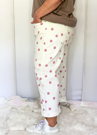 A person shown from the waist down wearing white pants with pink polka dots, a brown t-shirt, and white sneakers, standing on a white faux fur rug against a white backdrop.