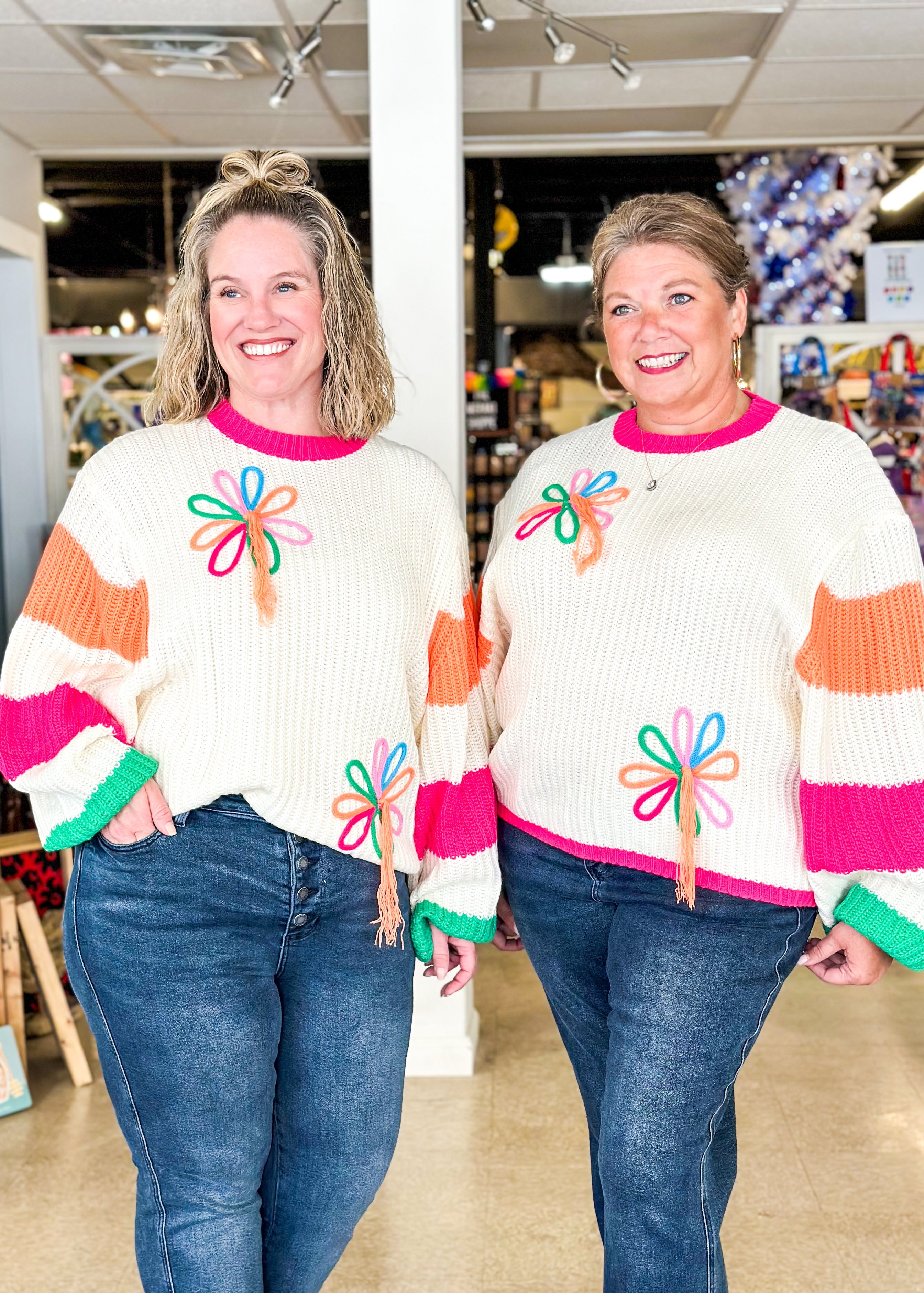 cream sweater with 2 bright flowers and orange tassels hanging from them, sleeves have wide orange and pink stripe, green around cuff and pink around bottom and neck